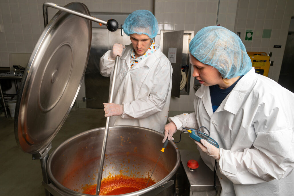 Two food scientists in white coats and hair nets stirring a large vat of red liquid